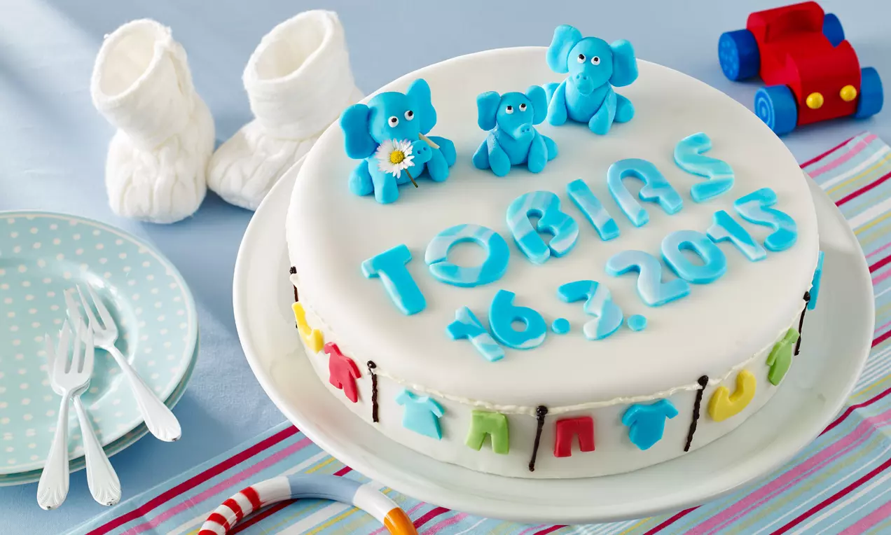 Runde Tauftorte mit weißem Fondant überzogen, oben blaue Schrift und Elefantenfiguren, seitlich bunte Kleidungsdekor auf weißem Teller