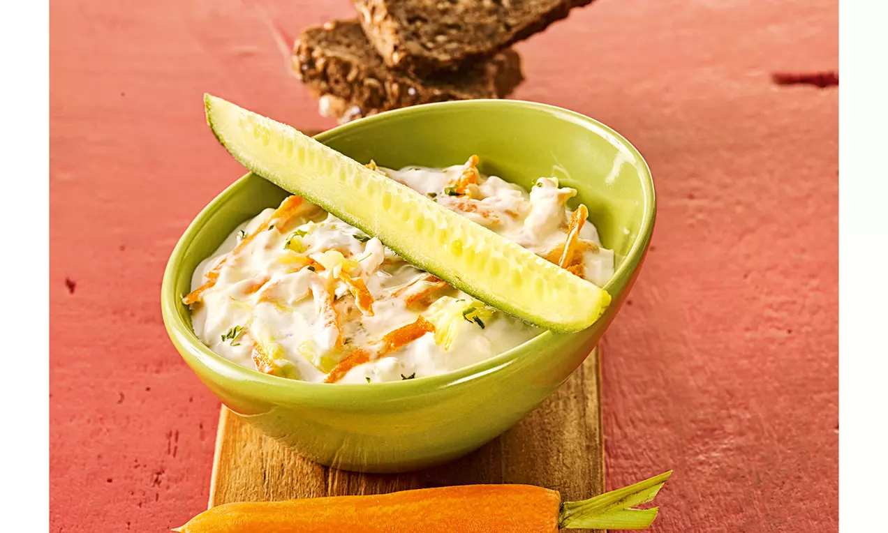 Cremiger weißer Dip mit orangefarbenen Möhrenstreifen und grünem Gurkenstift in einer grünen Schüssel auf Holzbrett mit Brot und Rohkost