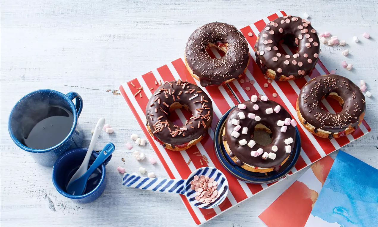 Donuts mit dunkler Schokoladenglasur, garniert mit bunten Streuseln, auf rot-weiß gestreifter Servierplatte neben blauen Tassen und Löffeln