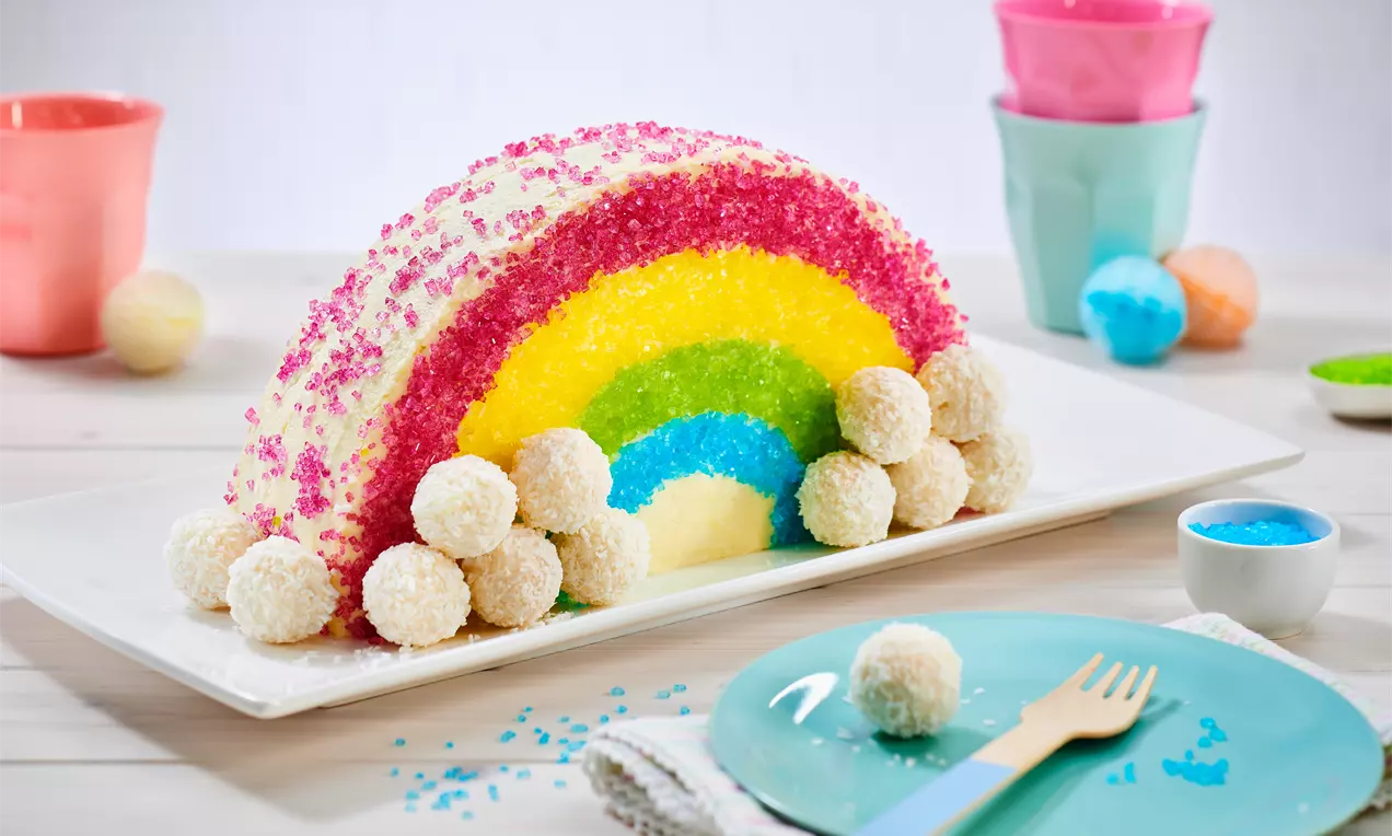 Bunter Regenbogenkuchen mit glänzendem Zuckerdekor und weißen Kokoskugeln auf rechteckiger Platte, umgeben von Pastellgeschirr