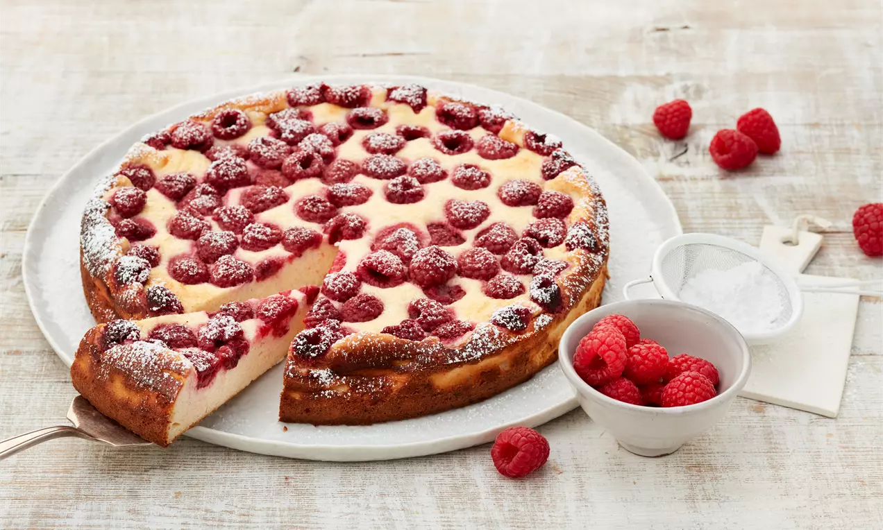Himbeer-Käsekuchen ohne Boden mit goldbrauner Kruste und saftigen Himbeeren, bestäubt mit Puderzucker auf weißem Teller mit frischen Himbeeren