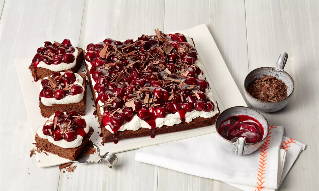 Rechteckiger Brownie mit dunkler Schokotextur, weißer Sahneschicht, Kirschkompott und geraspelter Schokolade auf einem weißen Teller
