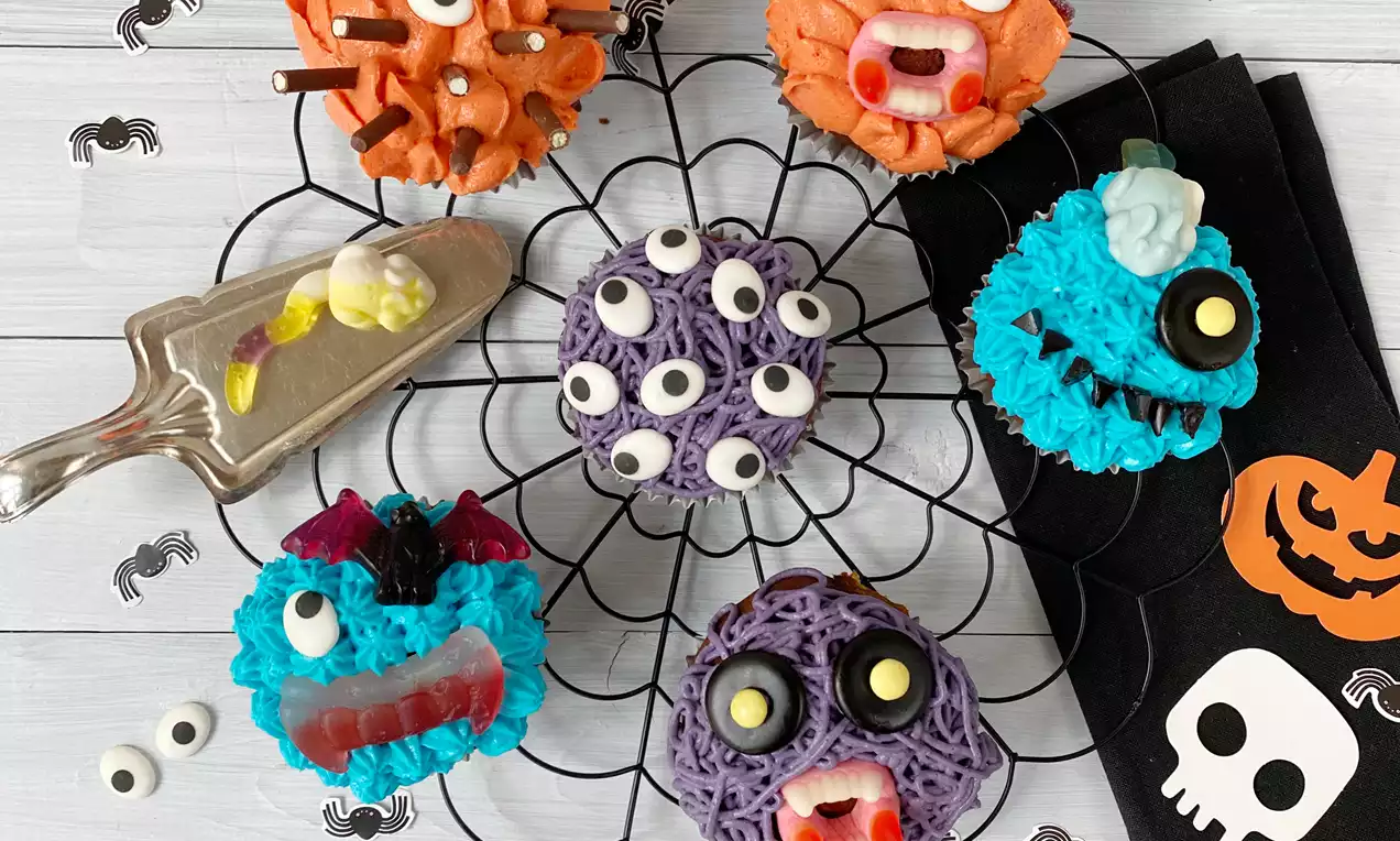 Bunte Halloween-Cupcakes in Monster-Designs mit leuchtendem Zuckerguss und Details wie Augen und Zähnen auf einem schwarzen Spinnennetz-Gitter