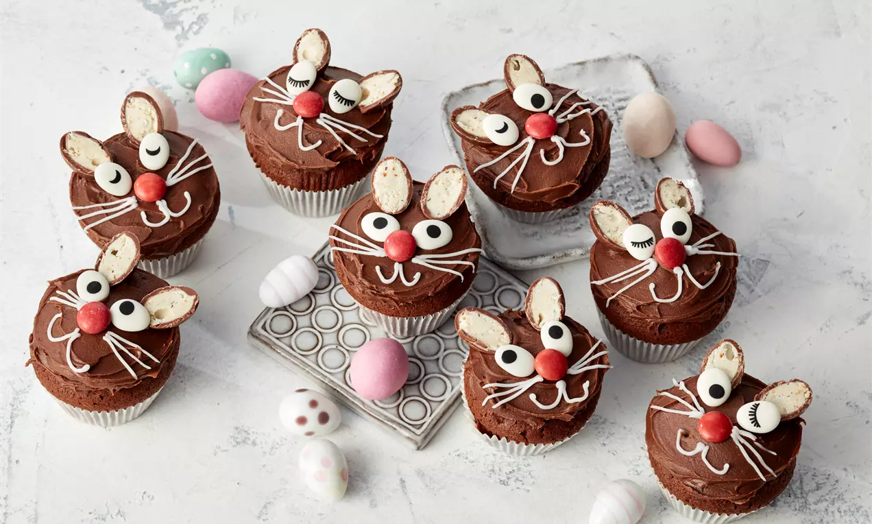 Oster-Muffins mit schokoladigem Teig, bunter Zuckerglasur, Mini-Marshmallows und Zuckerstreuseln, dekorativ in Pastell-Papierförmchen