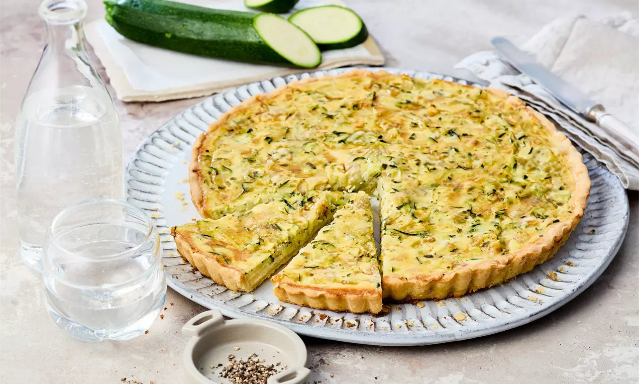 Vegetarische Zucchini-Quiche mit goldbrauner Kruste und grüner Füllung in einer geriffelten Backform, angerichtet mit frischen Zucchinischeiben
