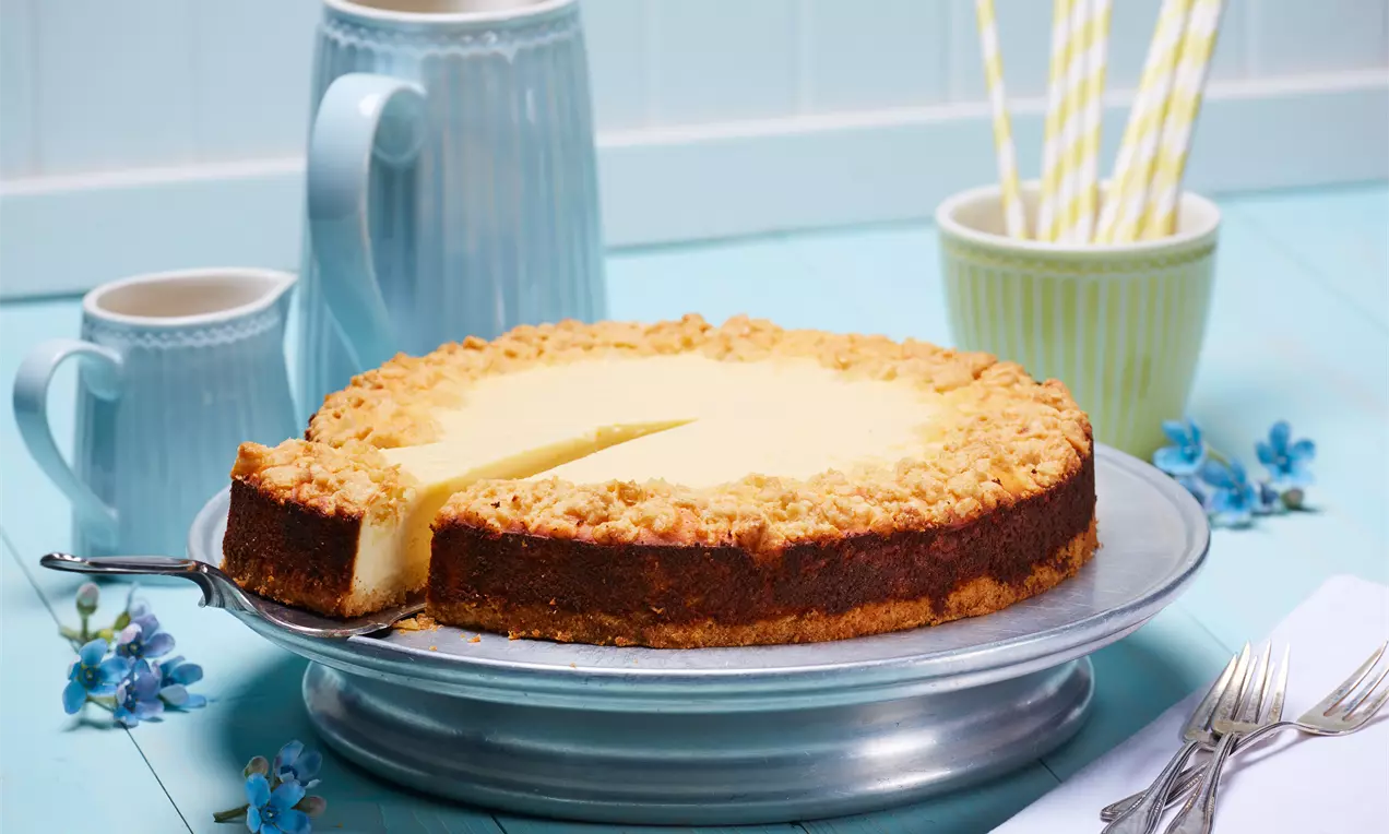 Käse-Streusel-Kuchen mit goldbrauner Kruste und hellgelber Füllung auf silberner Platte, umgeben von blauen Blüten und Tischgedeck