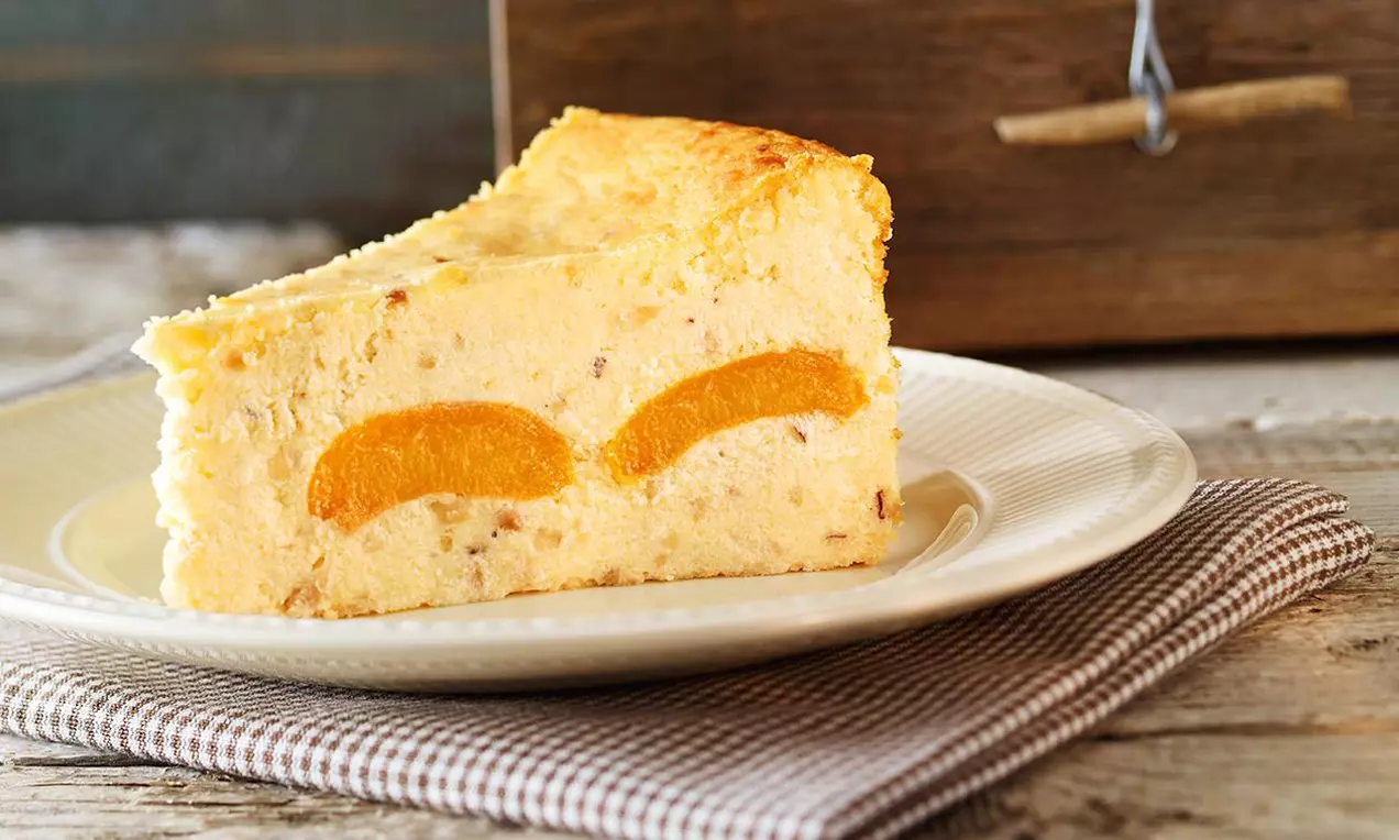 Ein Stück goldgelber Käsekuchen mit cremiger Textur und eingebetteten Aprikosenscheiben auf einem weißen Teller mit Stoffserviette drapiert