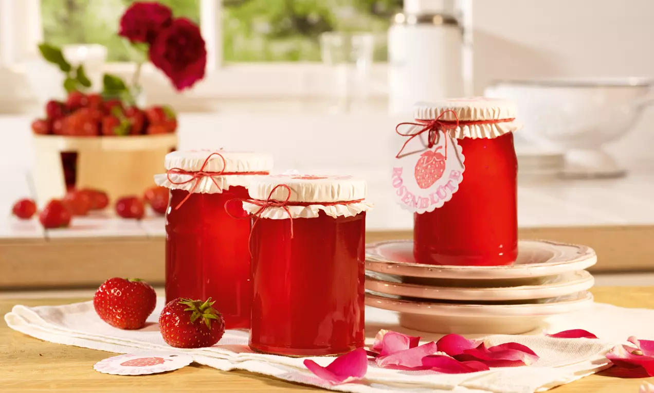 Gläser mit leuchtend rotem Erdbeergelee, mit Papierdeckeln und Schleifen gebunden, arrangiert auf weißen Tellern mit frischen Erdbeeren und Rosenblüten