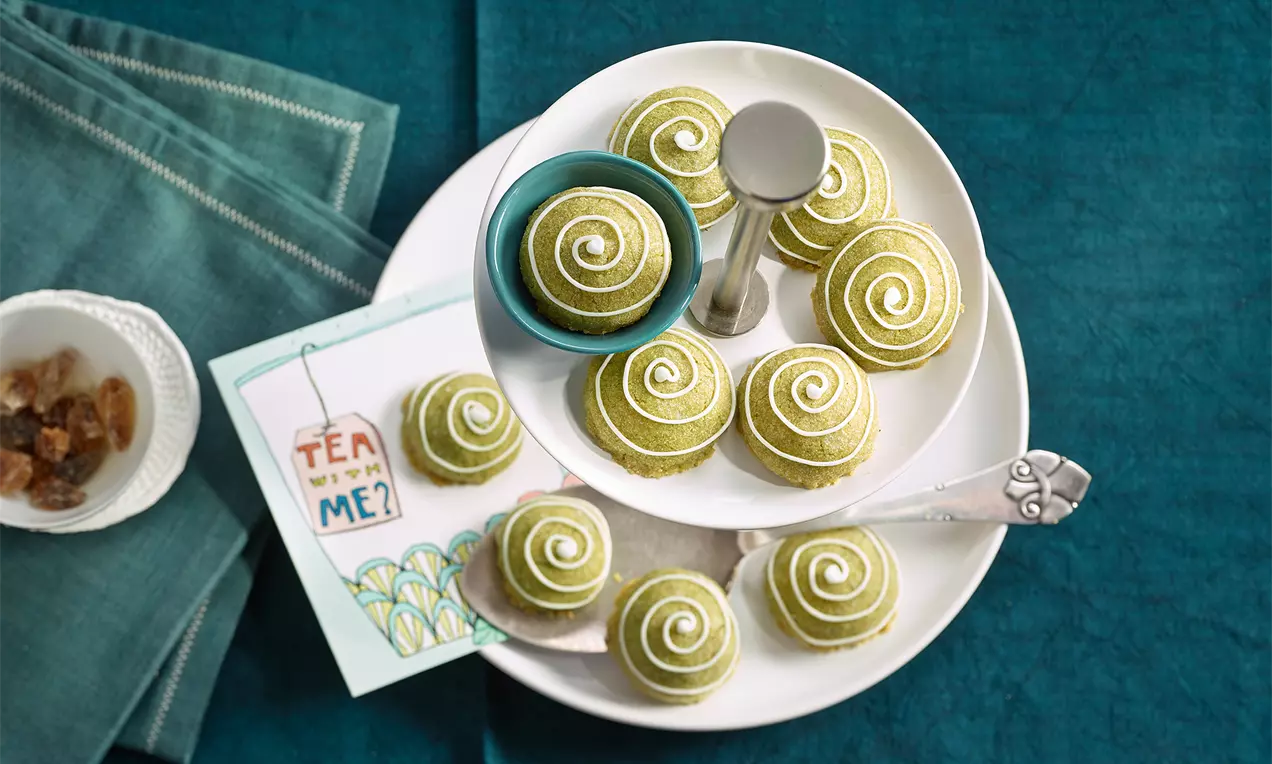 Matcha-Grün, spiralförmige Mürbeteigkekse mit weißem Zuckerdekor auf weißem Teller, Serviette und Teekarte auf petrolfarbenem Tisch