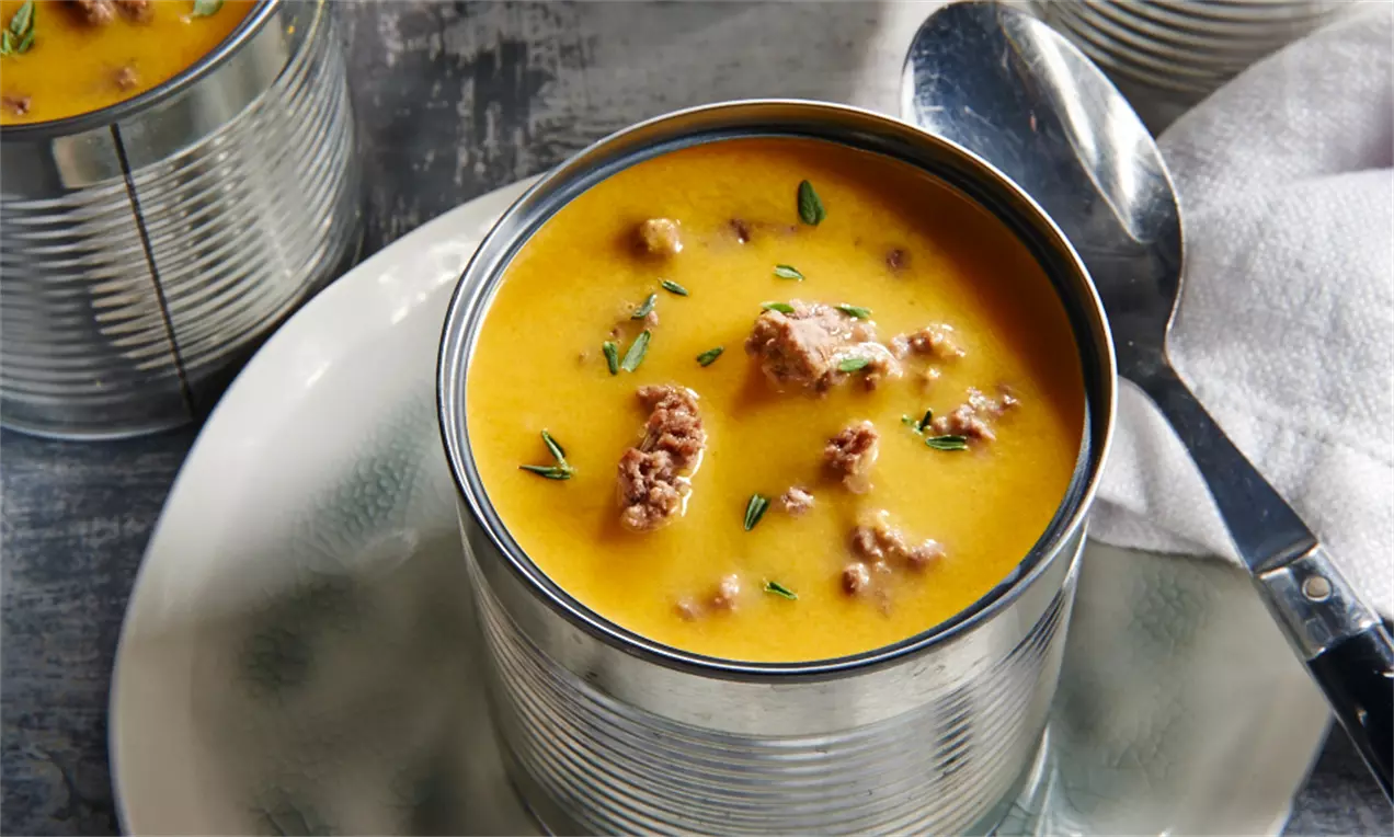 Kürbiscremesuppe mit orange-gelber Farbe, Stückchen Hackfleisch und grünen Kräutern in einer silberfarbenen Dose auf einem hellen Teller