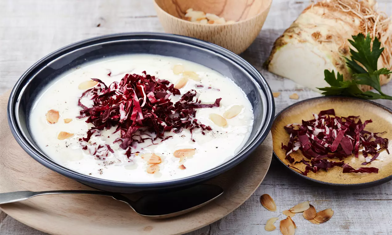 Cremige Kartoffelsuppe in graublauer Schale, bedeckt mit tiefrotem Radicchio und gerösteten Mandelsplittern, auf hellem Holzbrett angerichtet