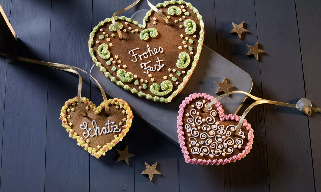 Drei braune Lebkuchenherzen mit bunter Zuckerdekoration und Schrift, mit goldener Schleife auf dunklem Holz und kleinen Deko-Sternen