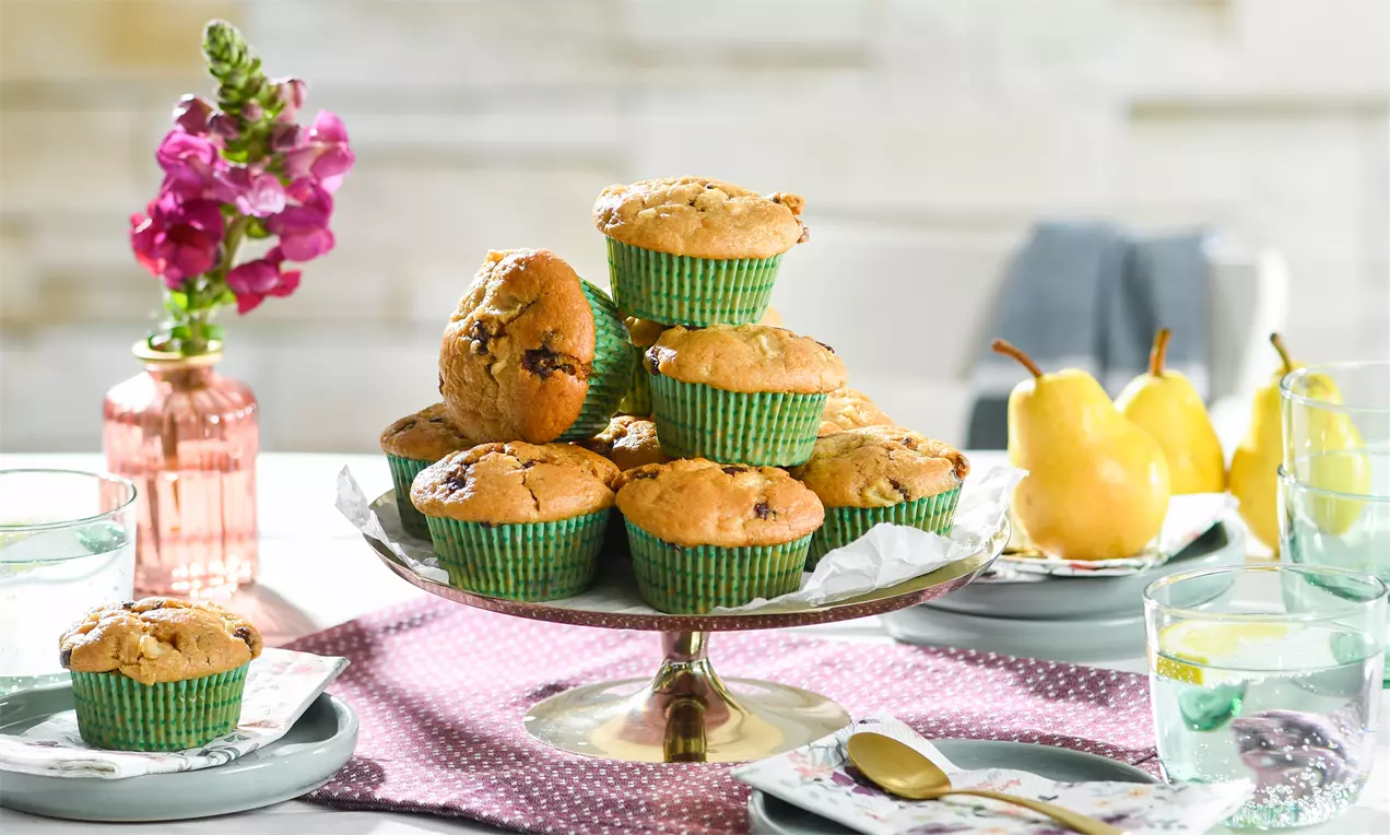 Goldbraune Birnenmuffins mit grünen Papierförmchen auf einer gläsernen Tortenplatte, umgeben von Birnen und Gläsern auf einem gedeckten Tisch