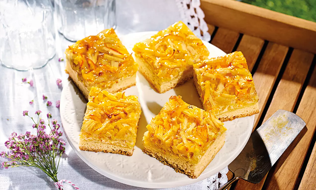 Quadratische Stücke eines goldgelben Apfel-Kürbis-Blechkuchens mit glänzender Oberfläche auf einem weißen Teller mit zarten Blüten dekoriert