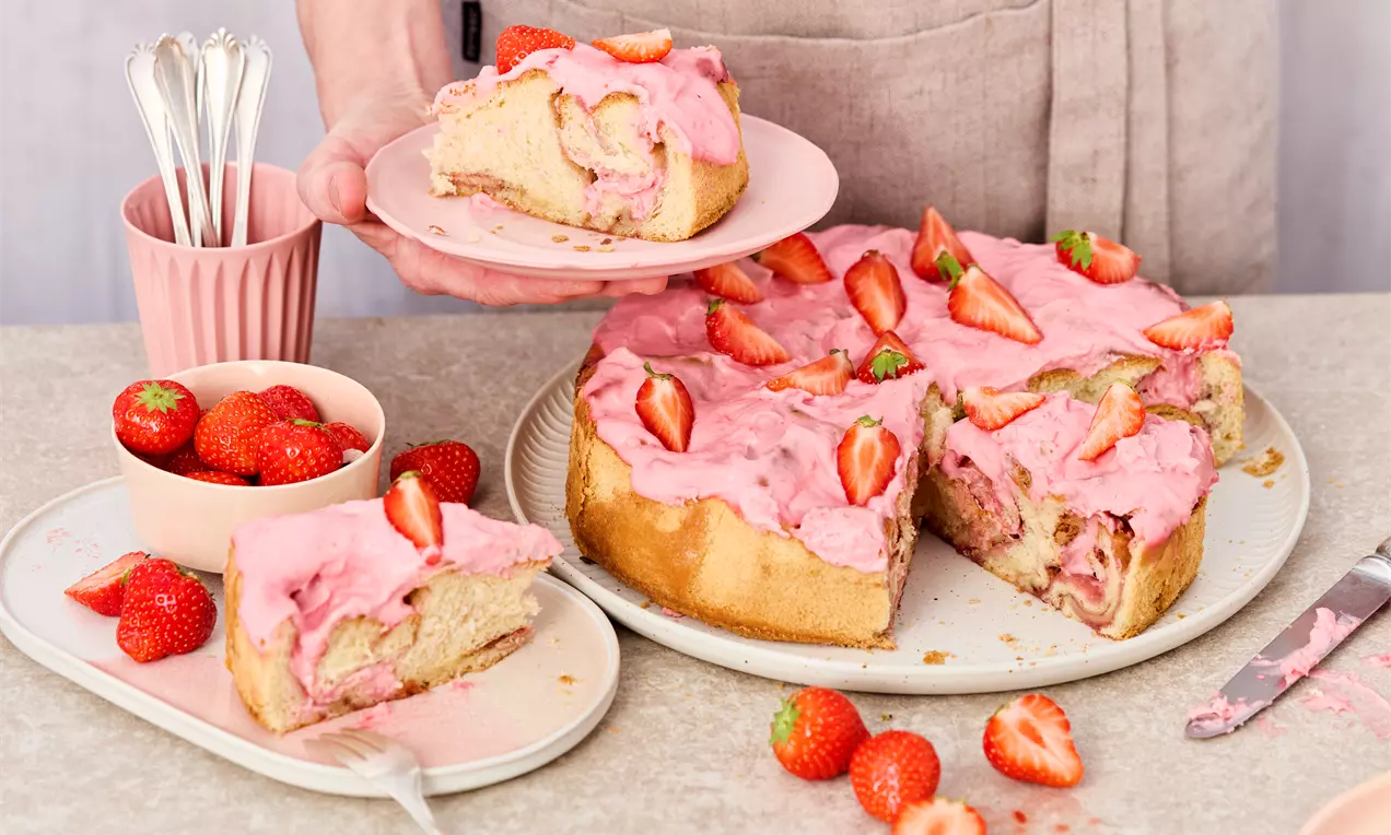 Erdbeer-Zimtschnecken in einer runden Form, überzogen mit rosa Erdbeercreme und dekoriert mit frischen Erdbeerstücken. Ein Stück liegt auf einem Teller, daneben eine Schale mit Erdbeeren und Besteck.