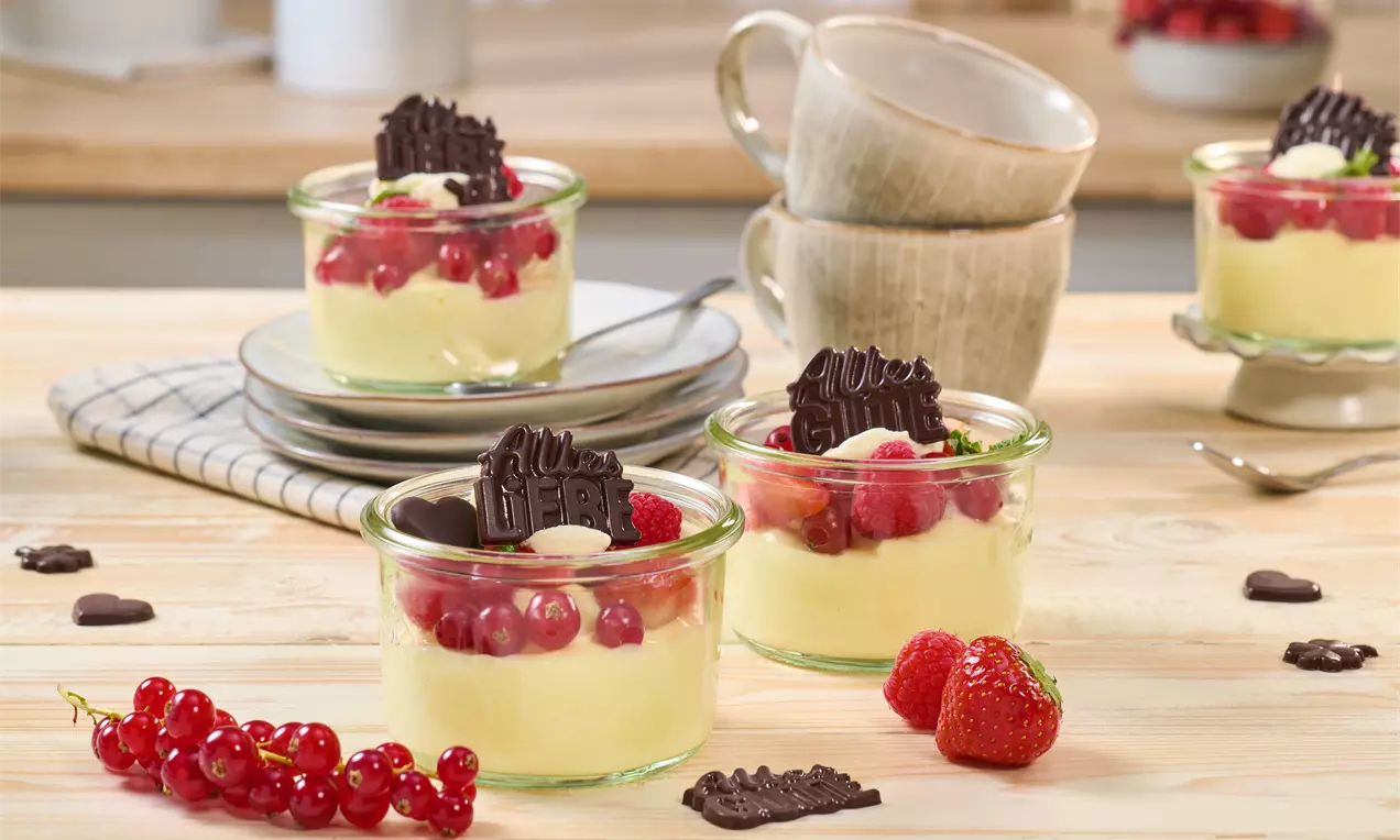 Geburtstagspudding in kleinen Gläsern, dekoriert mit roten Johannisbeeren, Erdbeeren und Schokoladenstücken. Im Hintergrund gestapelte Tassen, Löffel und weitere Puddinggläser auf einem Holztisch.