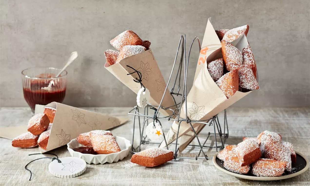Goldbraune, rechteckige Schmalzkuchen mit puderzuckerbestäubter Oberfläche in Papierhörnchen und auf Tellern, daneben rote Marmelade