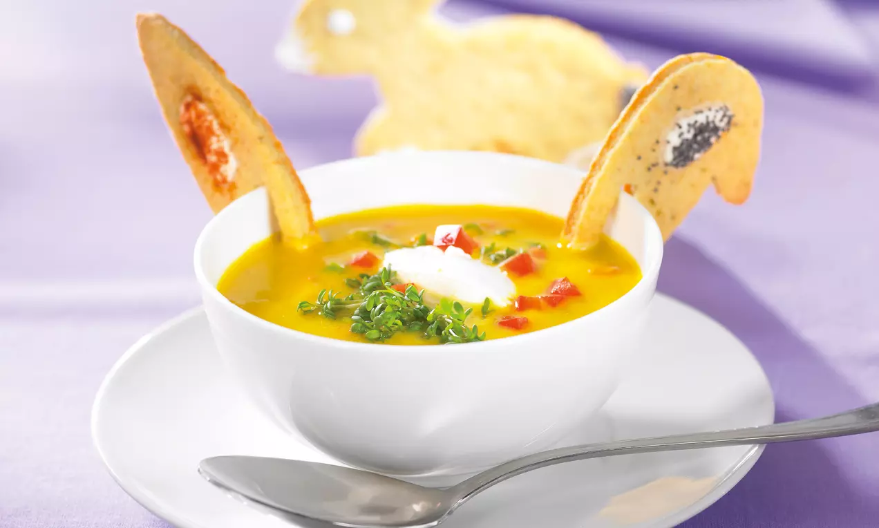 Leuchtend gelbe Suppe in weißer Schale mit Sahnehäubchen, roten Paprikawürfeln, grünem Schnittlauch und Hasen-Dekorkeksen auf hellem Untergrund