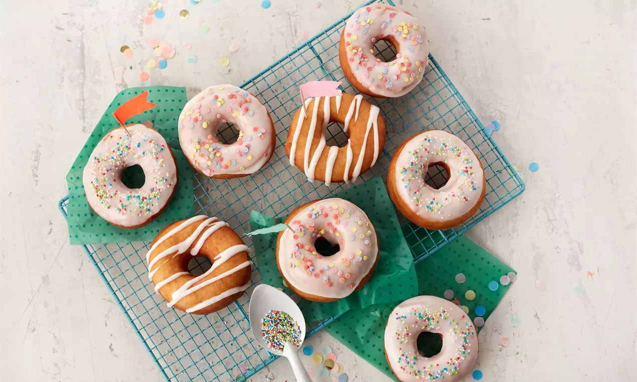 Donuts mit rosa und weiß glasierten Oberflächen, teils mit bunten Streuseln und Zuckerguss dekoriert, auf einem Gitter mit grünem Papier drapiert