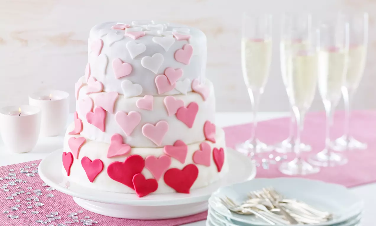 Dreistöckige weiße Hochzeitstorte mit rosa und roten Herzdekorationen auf einer Tortenplatte, daneben Sektgläser und rosa Tischdecke