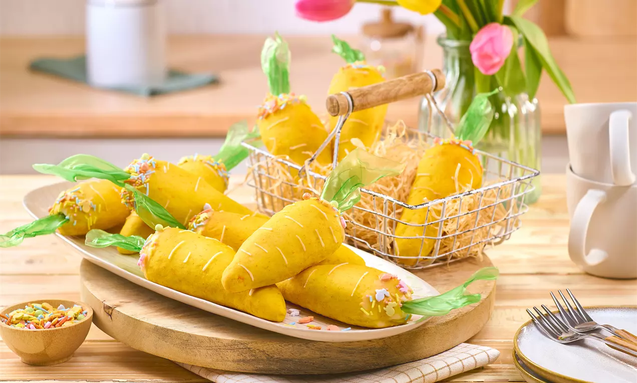 Karottenförmig geformte Hefeteig‑Milchbrötchen in leuchtendem Orange auf Platte und in Drahtkorb; dekoriert mit grünem Papier als Blattwerk und Blütenstreuseln, arrangiert auf Osterbrunch‑Tisch.