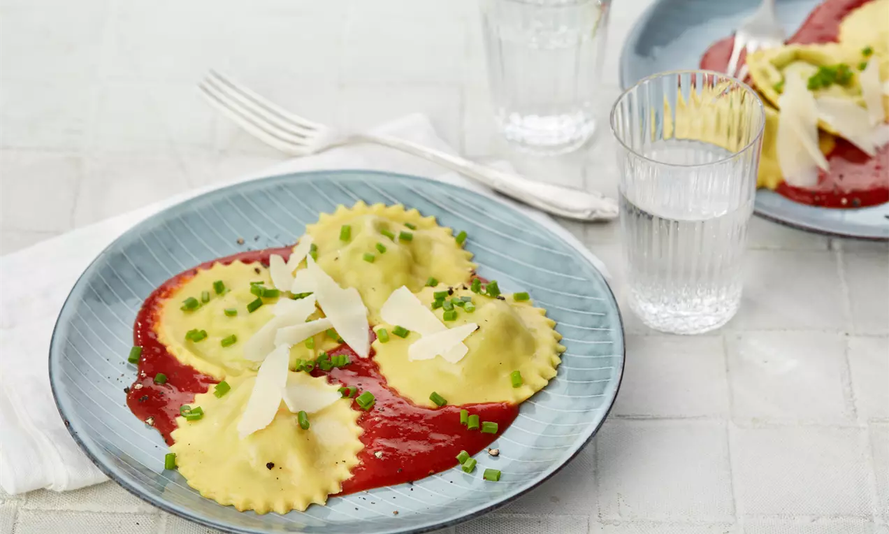 Gelblich matte Ravioli mit gewelltem Rand auf roter Tomatensauce, garniert mit hellen Parmesansplittern und grünen Schnittlauchringen