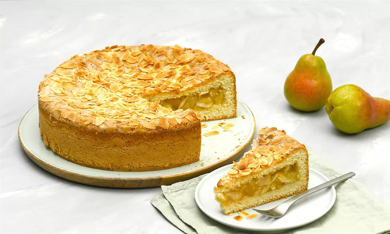 Gedeckter Birnenkuchen mit goldbraunem Mürbeteigboden, saftiger Birnenfüllung und gerösteten Mandelsplittern auf einem Teller mit frischen Birnen