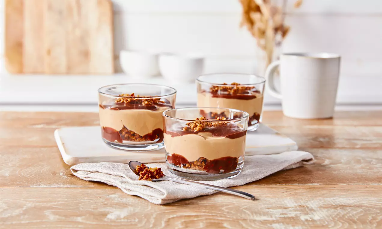 Drei Gläser mit geschichteter Karamellcreme, dunkler Soße und knusprigen Streuseln auf Holzbrett, mit Tuch und Löffel arrangiert