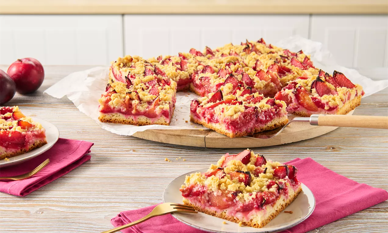 Pflaumenkuchen aus goldenem Quark-Öl-Teig mit saftiger roter Frucht und Streuseln auf Holzplatte, serviert mit Gabel auf rosa Stoffserviette
