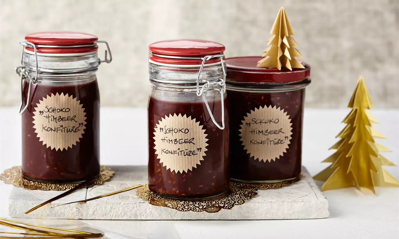 Schokoladige Himbeerkonfitüre im Glas mit leuchtend roten Himbeerstücken, dunkler Schokolade, frischen Beeren und Holzlöffel