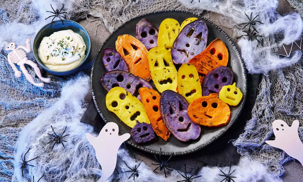 Bunte Kartoffelscheiben in Orange, Gelb und Lila mit geschnittenen Gesichtern auf schwarzem Teller, umgeben von Dekospinnen und Geistern