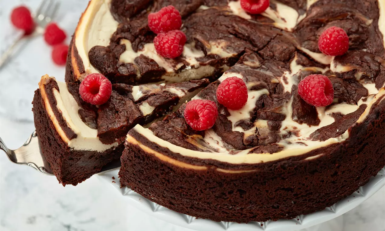 Brownie-Käsekuchen mit heller Marmorierung und saftiger, dunkler Kruste, dekoriert mit roten Himbeeren auf weißer Tortenplatte