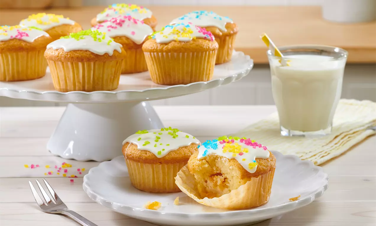 Goldgelbe Muffins mit weißem Zuckerguss und bunten Zuckerstreuseln auf weißer Keramik, daneben ein Glas Milch mit Strohhalm