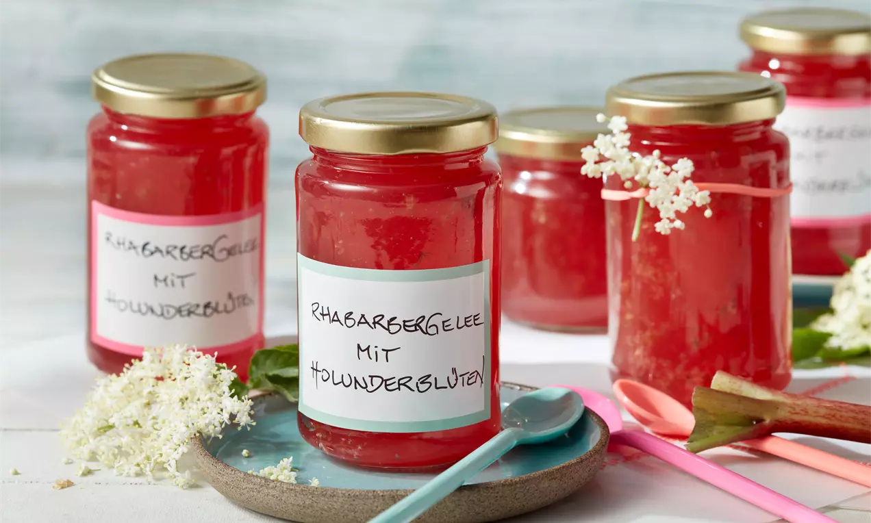Gläser mit leuchtend rotem Rhabarbergelee, goldenen Deckeln und weißen Etiketten, dekoriert mit Holunderblüten auf einem Holztisch