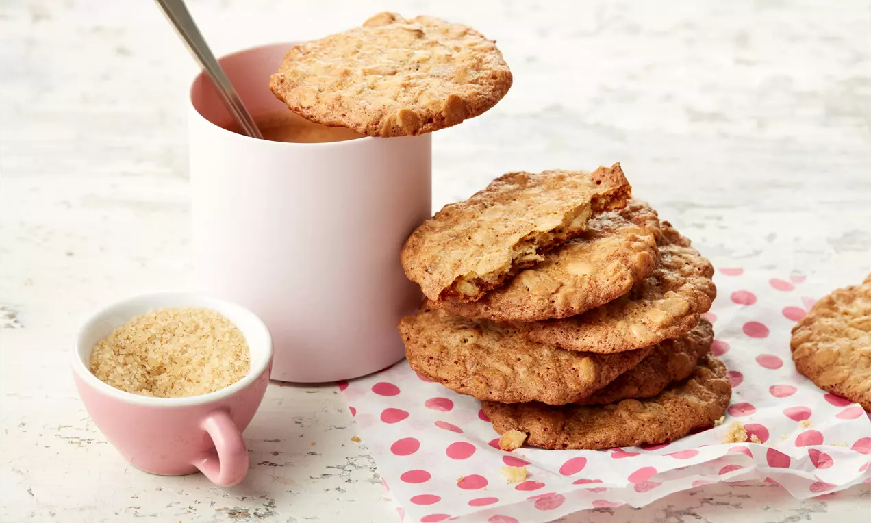 Flache, goldbraune Haferflockenplätzchen mit grober Textur, gestapelt auf gepunktetem Papier neben rosa Tasse und Schälchen mit Zucker