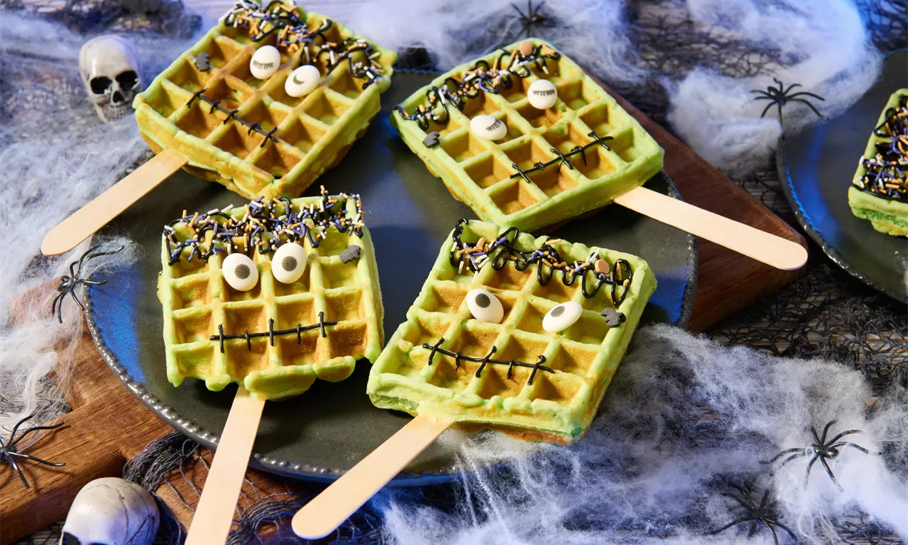Grüne rechteckige Waffeln am Holzstiel, mit Zuckerdekor verziert, Augen und schwarzen Linien auf einem Teller mit Spinnweben und Mini-Schädeln