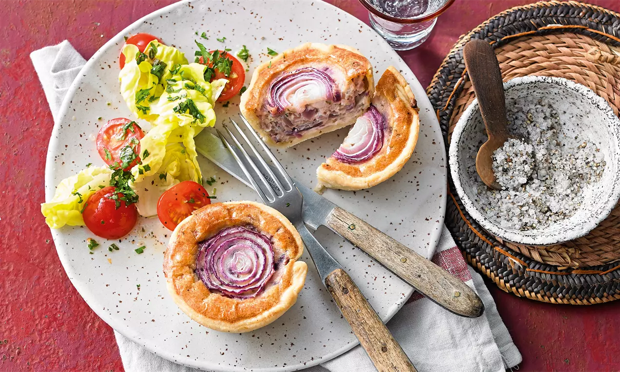 Zwiebelkuchen-Muffins mit goldbraunem Teigrand und lila Zwiebelringen auf einem weißen Teller mit Salat, Gabel und Messer angerichtet