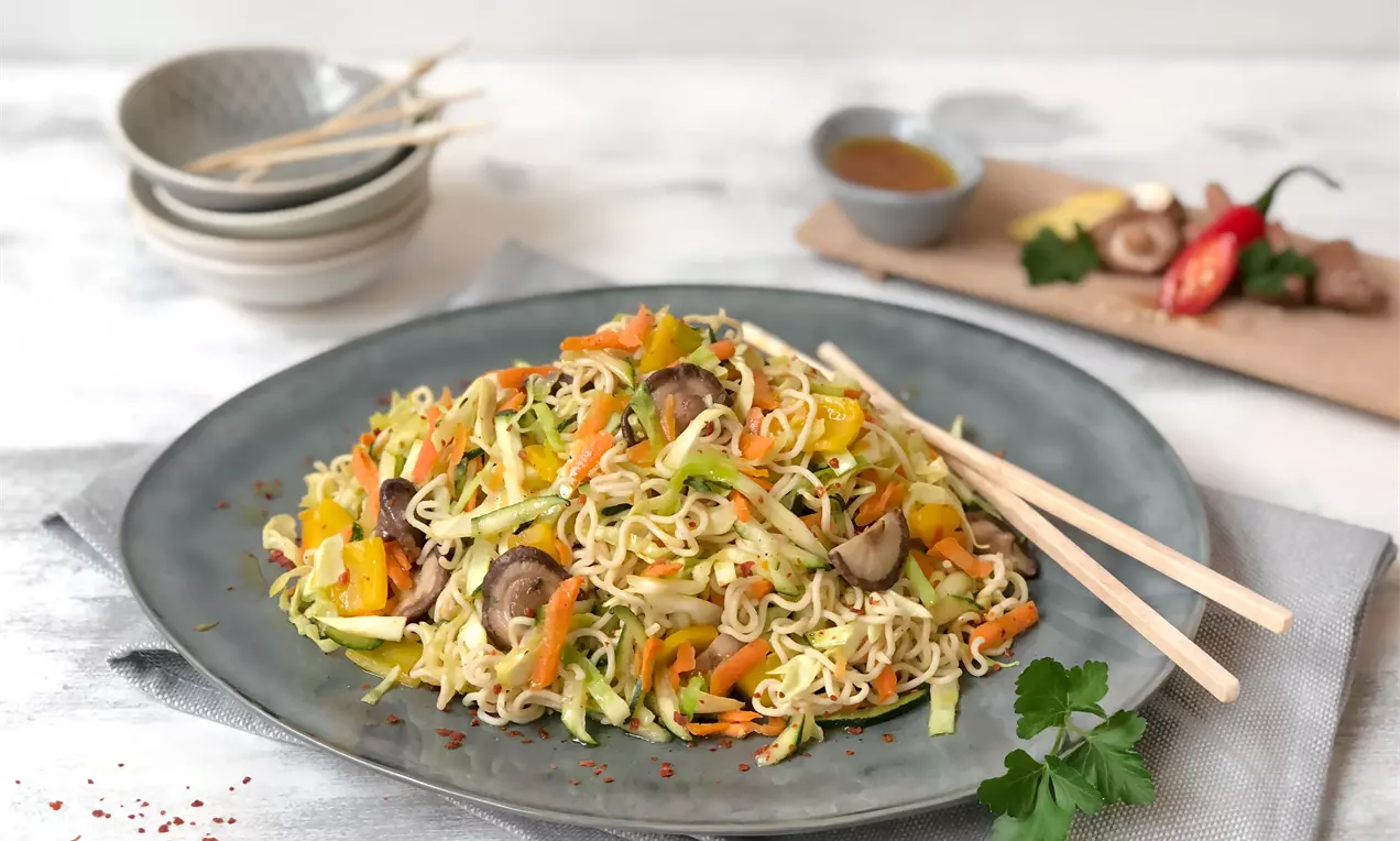 Asia-Salat aus goldgelben Nudeln, buntem Gemüse und Pilzen auf grauem Teller, angerichtet mit Holzstäbchen und Kräutergarnitur