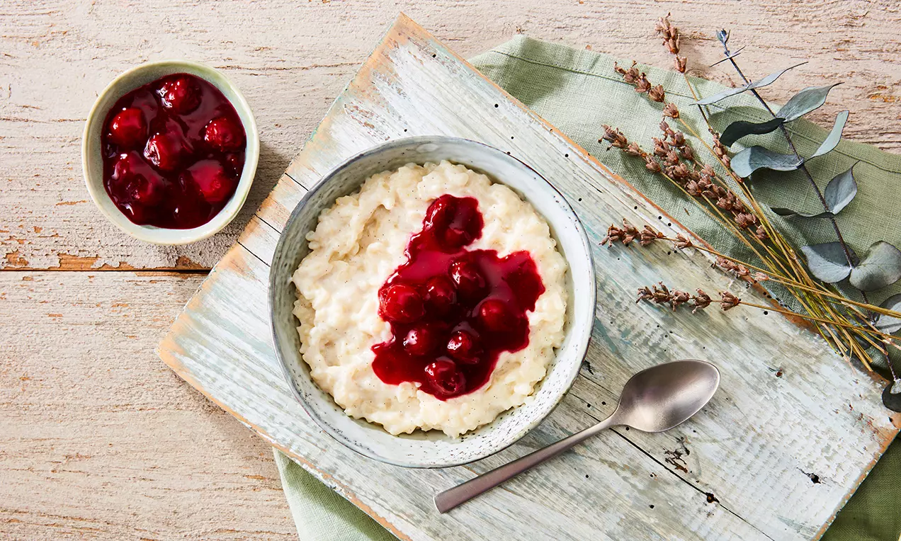 Cremiger weißer Milchreis mit glänzender roter Kirschgrütze in einer grauen Schale auf hellem Holzbrett mit grüner Serviette und Metalllöffel