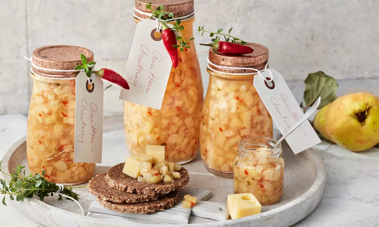 Gläser mit goldgelbem Quitten-Chutney, verziert mit Chilischoten und Kräutern, auf einem hellen Tablett mit Brot, Käsewürfeln und Quitte angerichtet