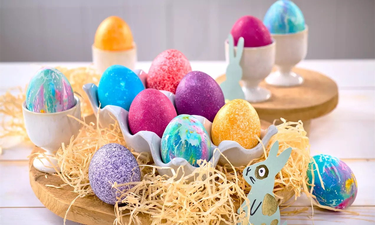 Bunte, marmorierte Ostereier in Stroh und Eierbechern auf einem hellen Tisch; mit Lebensmittelfarbe gefärbt und von kleinen dekorativen Hasenfiguren umgeben.