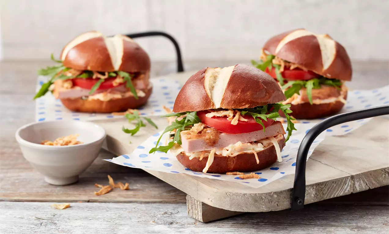 Runde Laugenbrötchen mit glatter, brauner Oberfläche, gefüllt mit rosa Leberkäse, grünem Salat, Tomatenscheiben und Röstzwiebeln