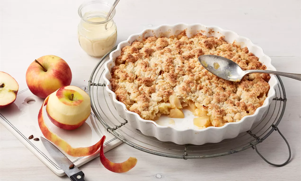 Apple-Crumble mit goldbraunen Streuseln und saftiger Zimt-Apfelfüllung in weißer Auflaufform, dazu Vanilleeis
