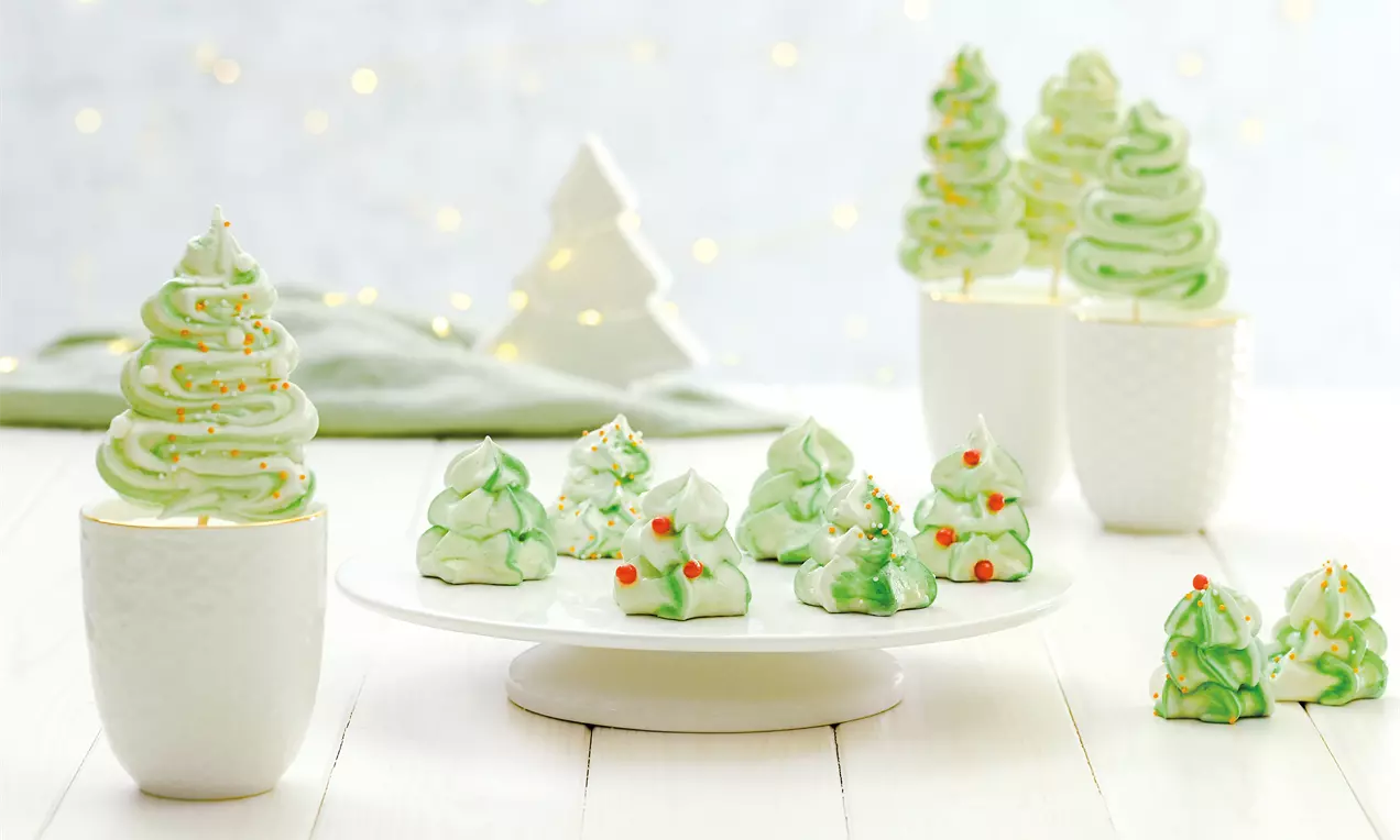Zarte grüne Baiser-Tannenbäume mit roten Zuckerperlen auf weißer Tortenplatte und in weißen Bechern, umgeben von dezenten Lichterpunkten