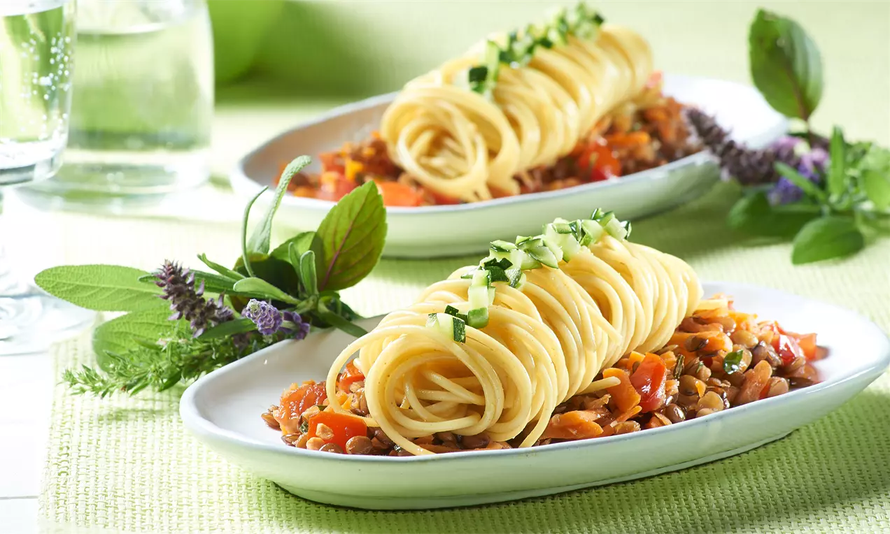 Dinkelspaghetti als goldgelbe Nester auf leuchtend orange-roter Möhren-Linsensoße, garniert mit grünen Kräutern auf weißem Teller