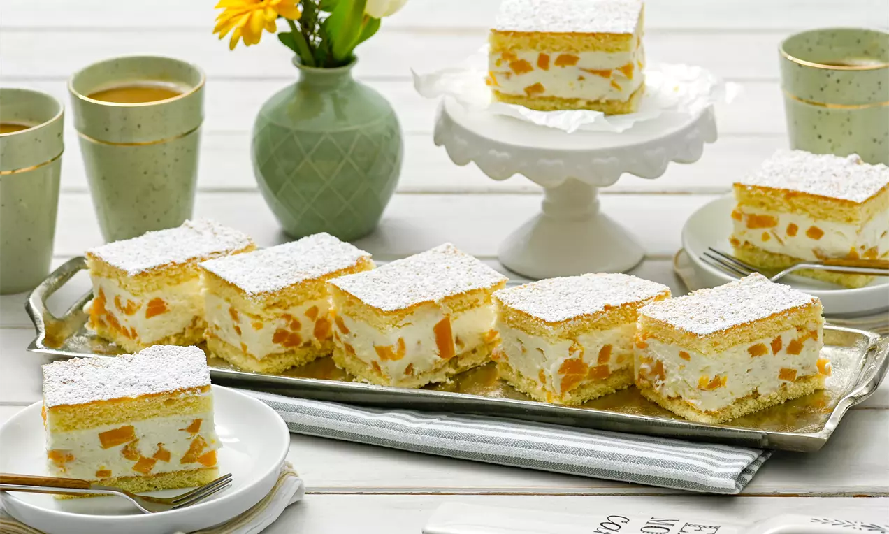 Pfirsich-Käse-Sahne-Schnitten mit heller Creme und saftigen Fruchtstückchen zwischen goldenen Biskuitlagen, mit feinem Puderzucker bestäubt auf einem Tablett angerichtet
