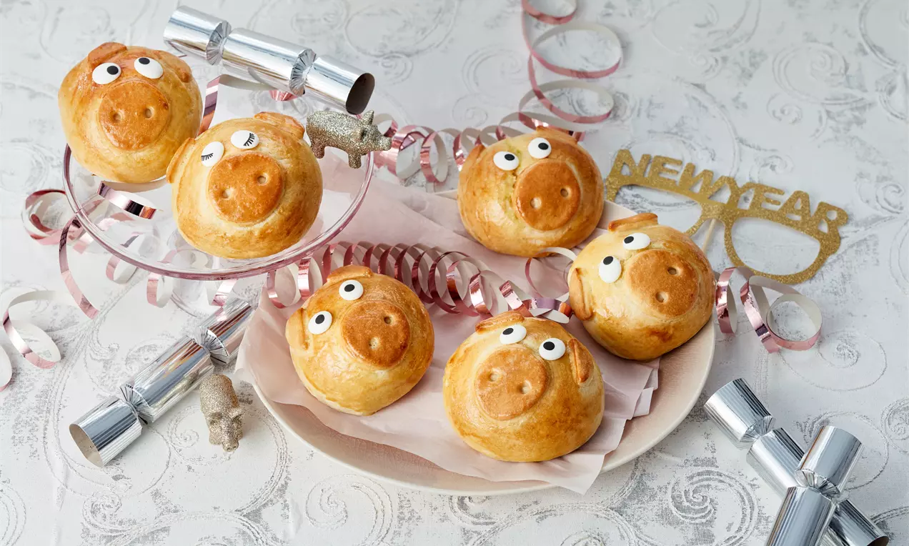 Goldbraune Hefeschweinchen mit glänzender Kruste, weißen Zuckergussaugen und rosa Dekor auf Tellern mit silbernen Partyaccessoires und Serpentinen