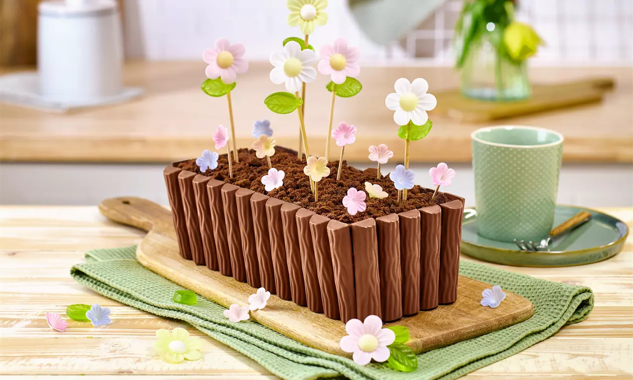 Kuchen in Form eines Blumenkastens mit Schokoriegel‑Rand und Kuchenkrümeln als Erde; dekoriert mit bunten Esspapierblumen auf Stielen, serviert auf Holzbrett und Stoffunterlage.