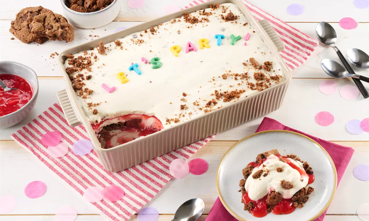 Cremiger Schichtdessert in einer rechteckigen Form mit buntem "LET'S PARTY!"-Schriftzug, dekoriert mit Cookie-Streuseln und rotem Fruchtkompott.
