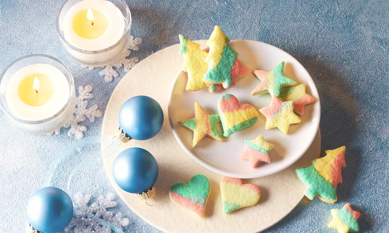 Bunte Adventsplätzchen in Stern- und Herzform mit rot-gelb-grünem Farbverlauf auf weißem Teller, neben blauen Kugeln und Kerzen drapiert